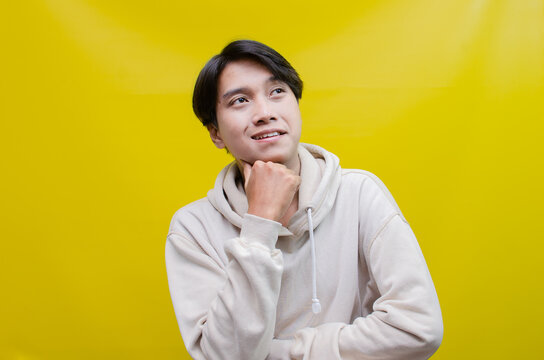 Thoughtful asian man in beige hoodie thinking and wondering with hand touching, rubbing head and face as thinking gesture isolated over yellow background