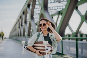 Fototapeta premium Young woman walking on city bridge with skateboard. Youth culture and commuting concept.