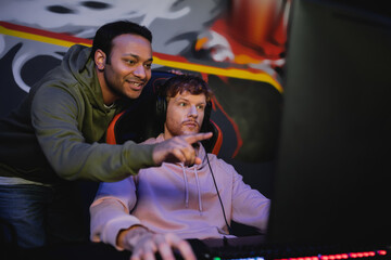 Cheerful indian man pointing at blurred computer monitor while friend in headphones playing video game in cyber club.