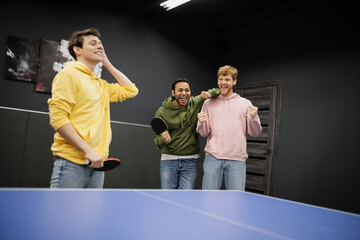 Excited interracial men standing near blurred friend with tennis racket in gaming club.