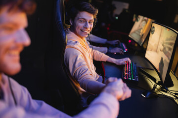 Fototapeta na wymiar Smiling man in headset shaking hand of blurred friend near computers in cyber club.