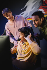 Young man wearing headphones near interracial friends and computer in gaming club.