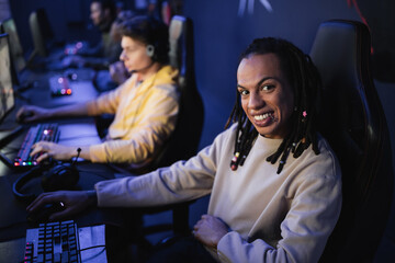 Multiracial gamer smiling at camera near blurred team in cyber club.