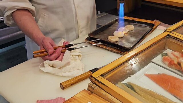 Professional japanese chef using hand-held burner burning to burn hotate scallops.
