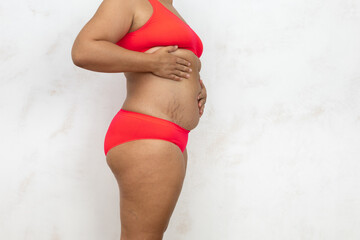 Overweight woman standing half turn massage belly by hands, free copy space, white background. Bare woman in red underwear with cellulite body and stretch marks. Plus size, feminism, self acceptance.