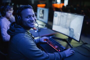 Smiling indian gamer in headphones looking at camera near computer in cyber club with lighting. © LIGHTFIELD STUDIOS