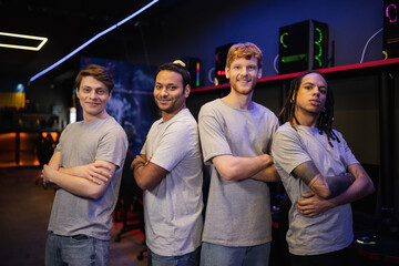 Smiling multiethnic gamer team in grey t-shirts crossing arms in gaming club.
