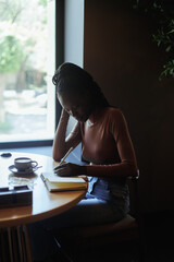 Portrait of young concentrated African-American woman with long dark braids wearing pink roll-neck sweater, sitting at beige table with cup of coffee, wooden tray in cafe, writing text in notebook. 