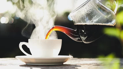 Poster Pouring hot black coffee to white coffee cup, mug with steaming smoke of coffee on old wooden table in morning nature outdoors, garden background. Hot Drink, Beverage Concept © iHumnoi