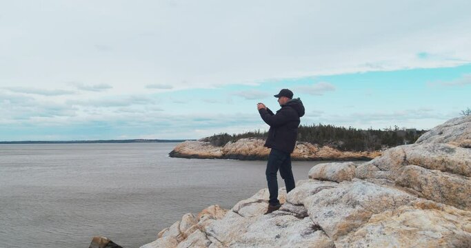 Man Tourist standing on the top of a rocky mountain, photographing the ocean with a smartphone and admiring the panoramic sunset against the backdrop of the Atlantic Ocean.