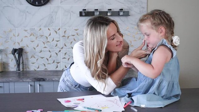 A young blonde woman with long hair draws pictures with a charming girl of 4 years old in the kitchen at home. A happy family. Mother and daughter painting together. An adult woman helps a girl.