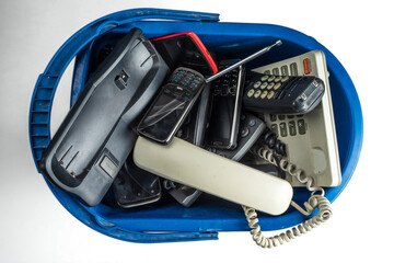 Fototapeta premium Old desk phones, cordless phone, cell phones, and smartphones in a trash can isolated on a white background.