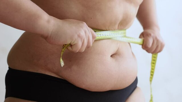 Close-up of unrecognizable buxom woman wearing black underwear, tightening flabby waist with yellow measuring tape, turning around on white background. Weight control, plastic surgery, liposuction. 
