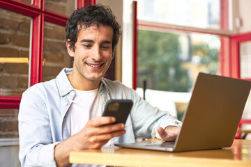 Young man happily multitasking on laptop and phone in cozy cafe.