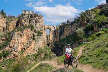 nice, active senior woman riding her electric mountain bike below the famous New Bridge of Ronda, Andalusia, Spain