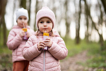 Two sisters drink juice using straws at forest, happy child moments.