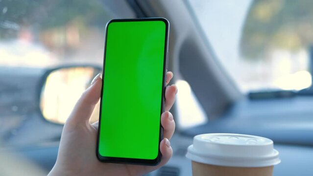 A hand holding a smartphone with green blank screen and coffee in the car for direction, massage, location, business. Woman sits in modern car and works on smartphone - closeup. Chroma key.
