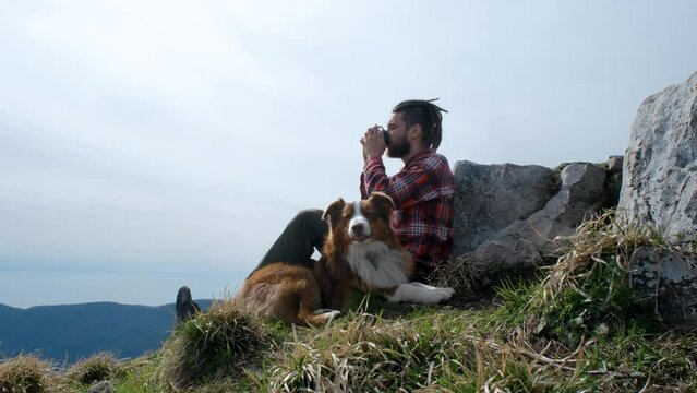 Travel concept. Caucasian man with dreadlocks sitting on top of mountain and drinking hot tea. Guy traveling with his dog, n Australian Shepherd. Rest during the hike.