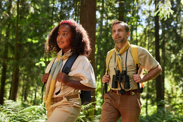 Waist up portrait of cute girl scout hiking in forest with adult leader scene lit by sunlight