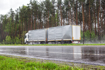 a road train with a semi-trailer transports cargo on a road slippery from rain in bad cloudy weather against the backdrop of a forest, industry