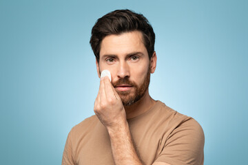 Naklejka premium Handsome bearded man applying cleansing lotion on his face, using cotton pad, looking at camera, blue studio background