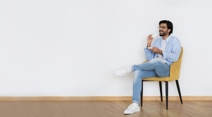 Positive young islamic male in casual and glasses sits on armchair have rest, enjoys spare time, drinking coffee or tea