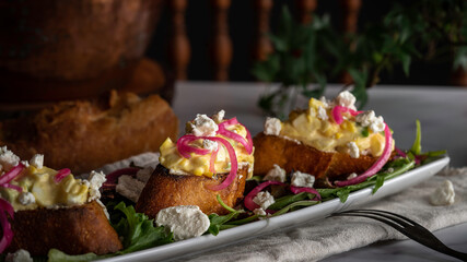 Creamy egg salad tops grilled slices of baguette on a bed of arugula. Garnished with pickled red onions and crumbled goat cheese.