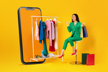 Mobile shopping offer. Shopaholic woman pointing at clothes on clothing rail in huge cellphone over...