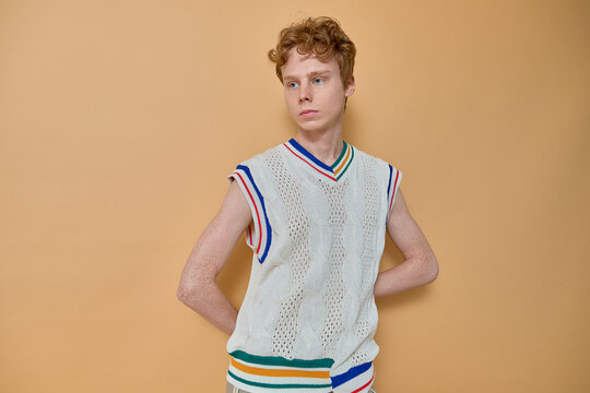Young focused student with red hair and freckles over his body wearing knitted vest with colored stripes standing with his hands behind back looking away and posing for photo on yellow wall.  - Powered by Adobe