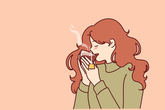 Woman drinks hot tea from transparent glass, enjoying taste of warming drink in cold winter weather. Young girl in sweater feels cozy and drinks tea drink take break to rest from work 