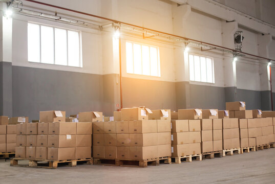 Pallets With Ready-made Cardboard Boxes Are Stored In A Warehouse. Concept Production For The Production Of Cardboard Boxes From Corrugated Cardboard. Rays Of Sunset Through The Window Indoors, 