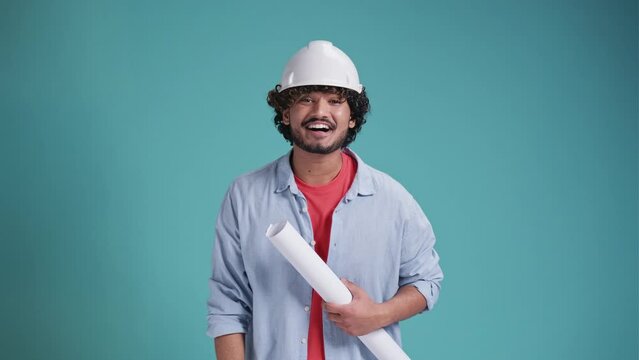 Young indian or arabic engineer standing holding helmet, blueprint and wearing ear muff standing happy delighted isolate on a blue background.