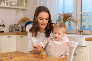 Happy family at home. Mother feeding baby in kitchen. Little boy with messy funny face eats healthy food. Child learns eat by himself holding spoon. Woman mom giving food to kid son. Self feeding