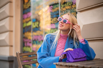 Happy smiling fashionable woman wearing trendy pink rectangular sunglasses, turtleneck, blue coat, with quilted purple bag, posing outdoors. Copy, empty space for text
