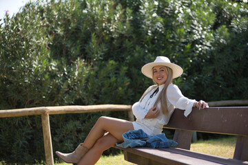 Young, beautiful, blonde woman with green eyes, wearing white shirt and hat, miniskirt, sitting on a wooden bench, smiling, relaxed and happy. Concept beauty, fashion, trend, relax, happiness.