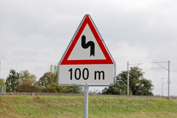 swiss traffic sign: double bend starting to the left. in 100 meters. cloudy during the day without people. drive slowly situation can be dangerous
