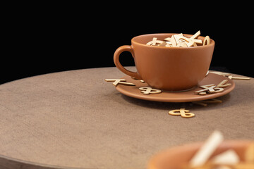 A ceramic cup on a plate overflowing with wooden letters on a wooden table in front of a black background