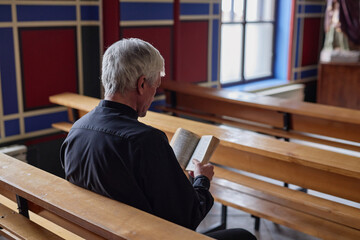 Rear view of senior priest reading Bible during praying while sitting on bench in church