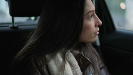 Fototapeta na wymiar One pensive young woman sitting in car backseat looking out window. Thoughtful expression of a female passenger riding taxi