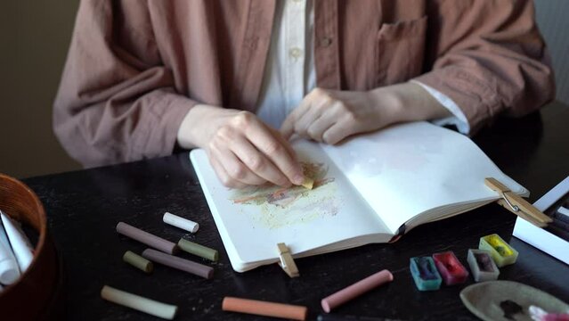 Woman painter artist drawing picture in album nature house using pastel chalks sitting at table, hands close-up. Girl art student practicing painting skills doing homework. Paint art classes concept.