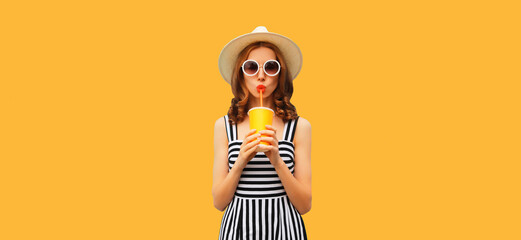 Summer portrait of beautiful young woman drinking fresh juice wearing straw round hat, striped dress on orange background