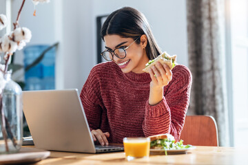 Beautiful young woman working with laptop while having healthy sandwich for breakfast at home