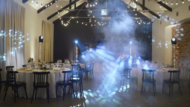 Decorated banquet hall in old stone building with round set tables and black chairs with main table in center. There is smoke machine and professional stage lighting with garlands and rotating beams.
