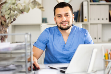 Portrait of positive man doctor in surgical scrubs sitting at working table in his office and using laptop.