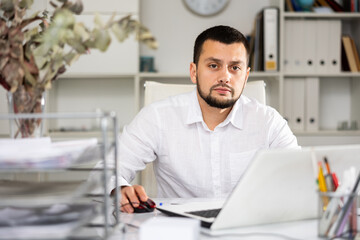 Fototapeta premium Male bookkeeper sitting at table and using laptop while doing his job in office.