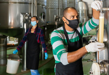 Portrait of male winery worker in mask controlling winemaking process, checking wine near metal tanks for wine fermentation
