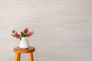 Red callistemon bottle brush flowers in white jug on wooden stool against white painted wall...