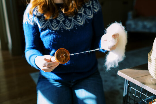 Woman in bright blue sweater in her living room and hand spinning yarn
