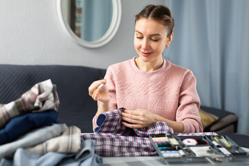 Smiling young woman sewing on button on shirt at living room - obrazy, fototapety, plakaty