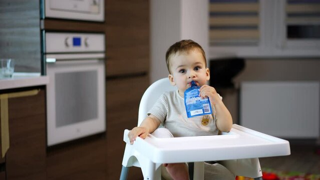 Cute Caucasian baby boy sits in a high chair. Toddler is holding a fruit puree pack in his mouth and smiles adorably to the camera. Blurred backdrop.